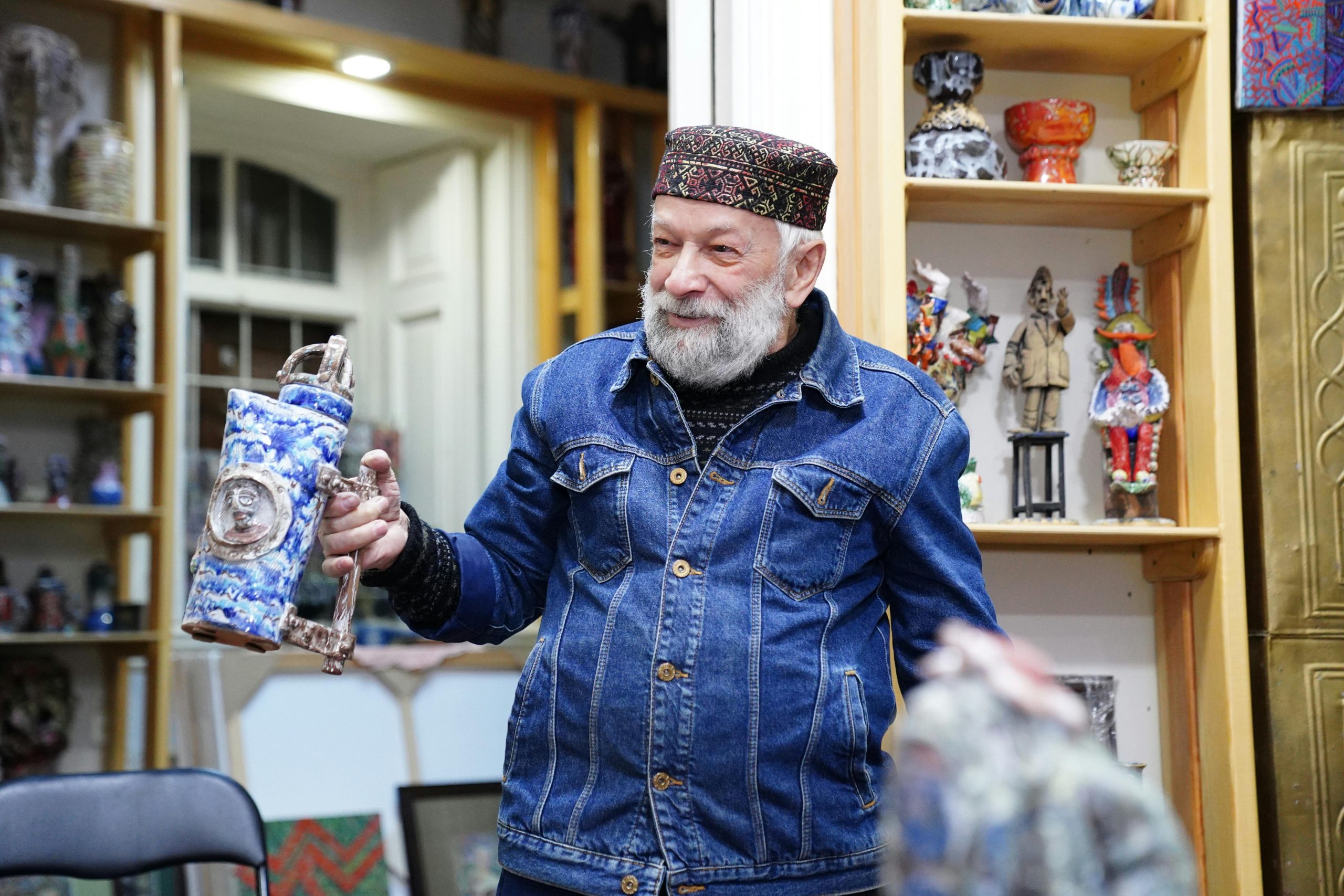 Elderly man in traditional Azerbaijani attire holding artistic pottery, surrounded by vibrant crafts indoors.