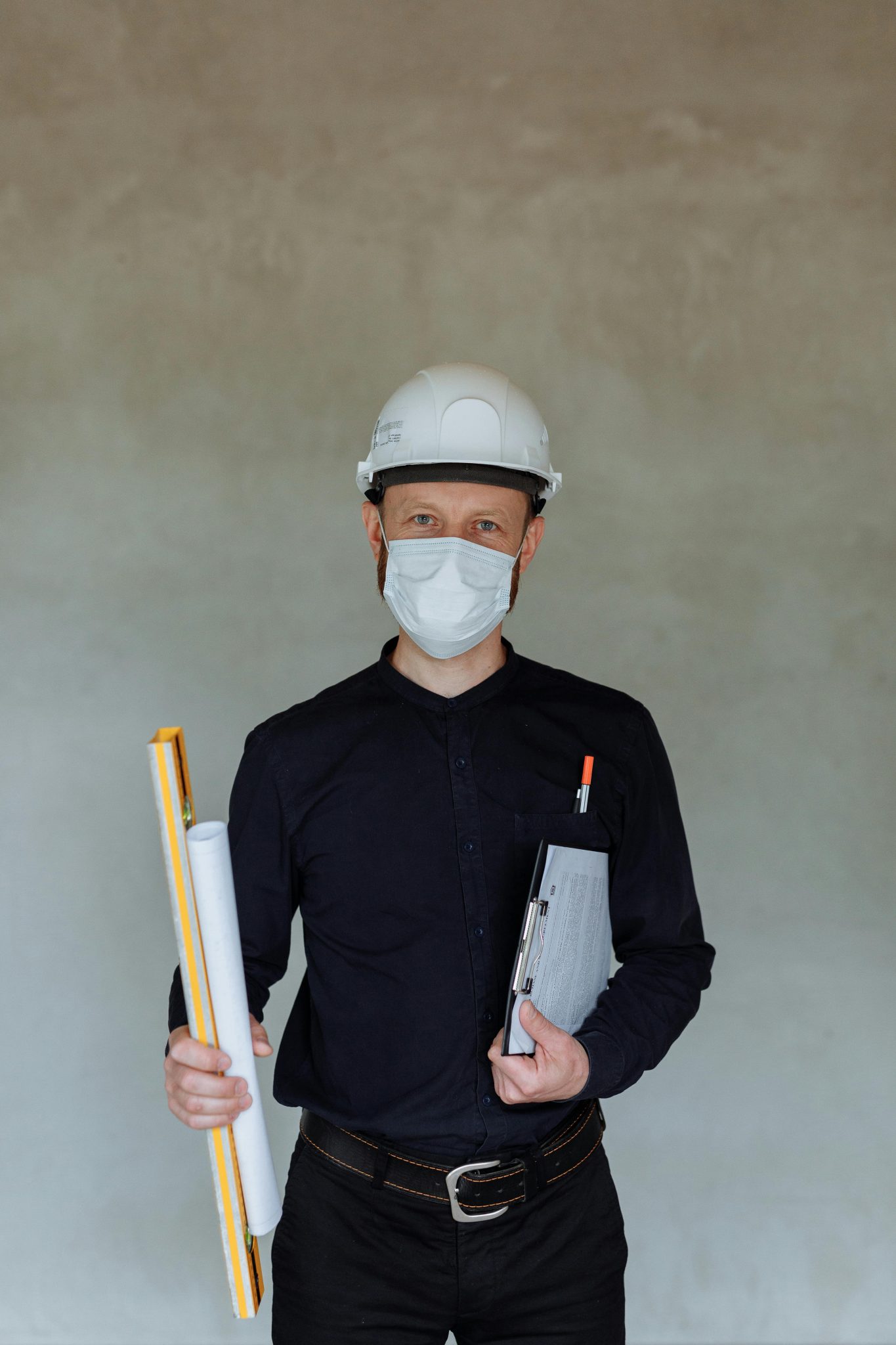 Architect wearing a hard hat and face mask, holding blueprints indoors.