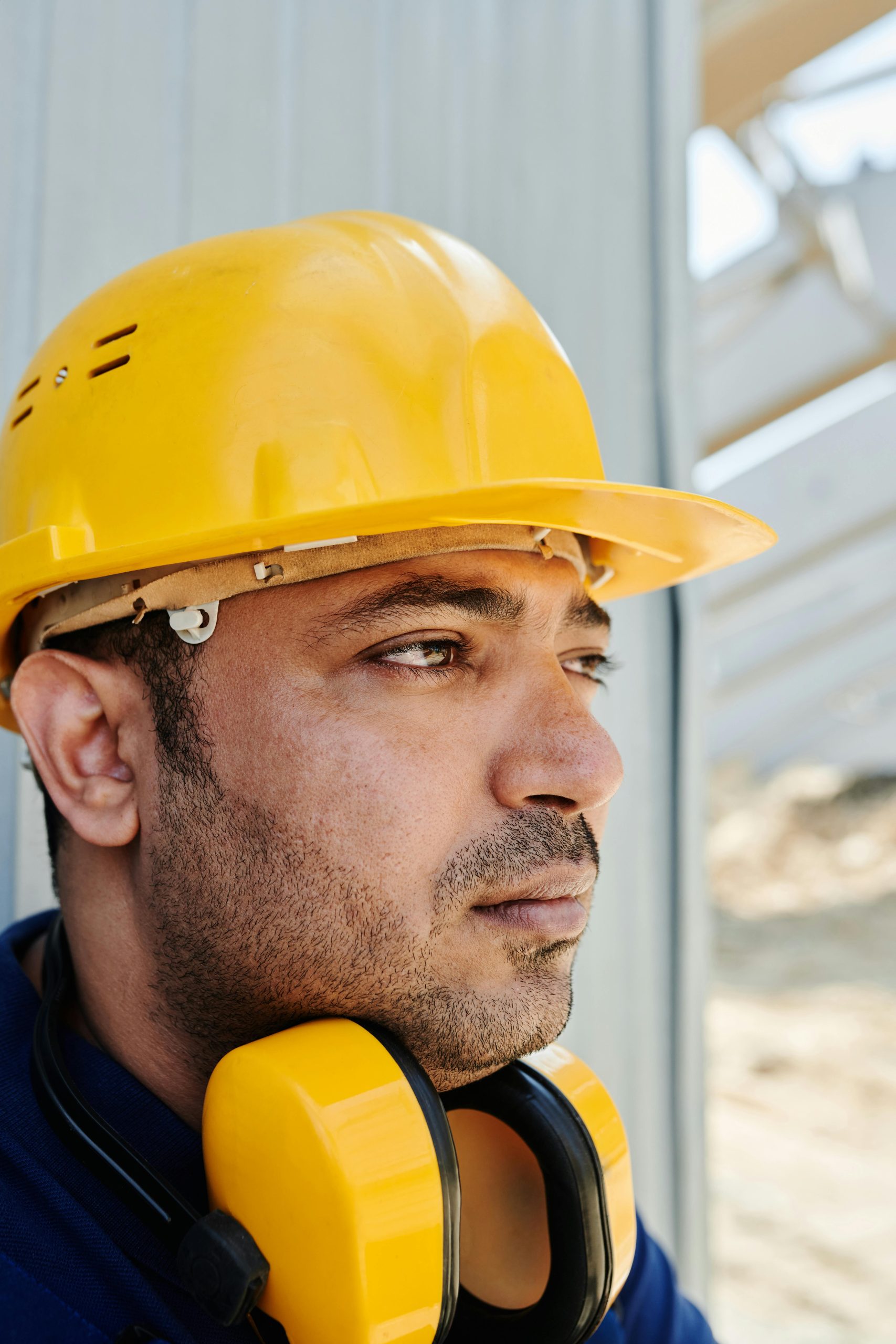 Close-up of a focused construction worker wearing a yellow hard hat and safety earmuffs outdoors.
